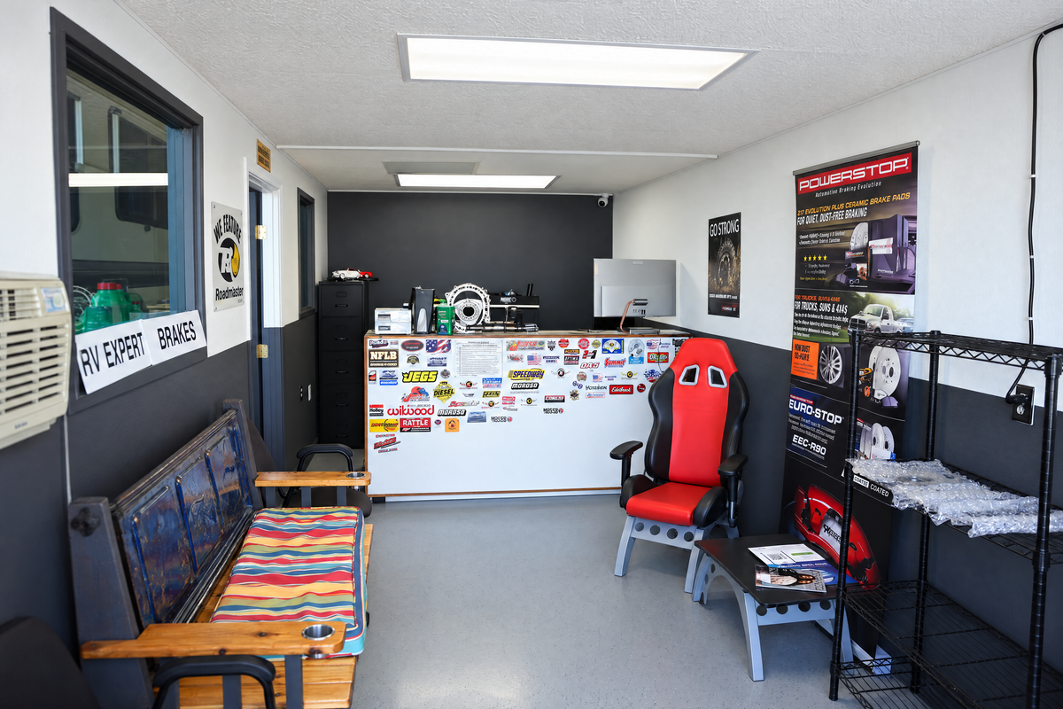 The waiting area: a counter covered in parts-brand stickers, a red racing-seat chair, a pickup-tailgate bench and brake displays