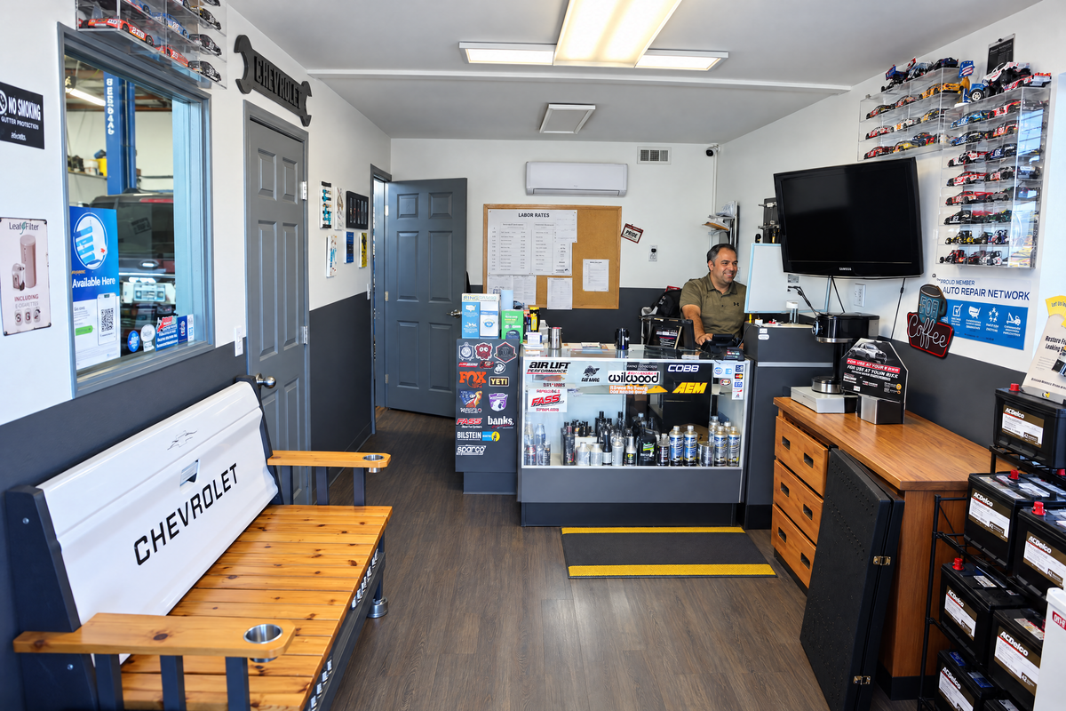 The front counter, with a man standing behind it, a parts display case, model cars on the wall and a Chevrolet tailgate bench in front