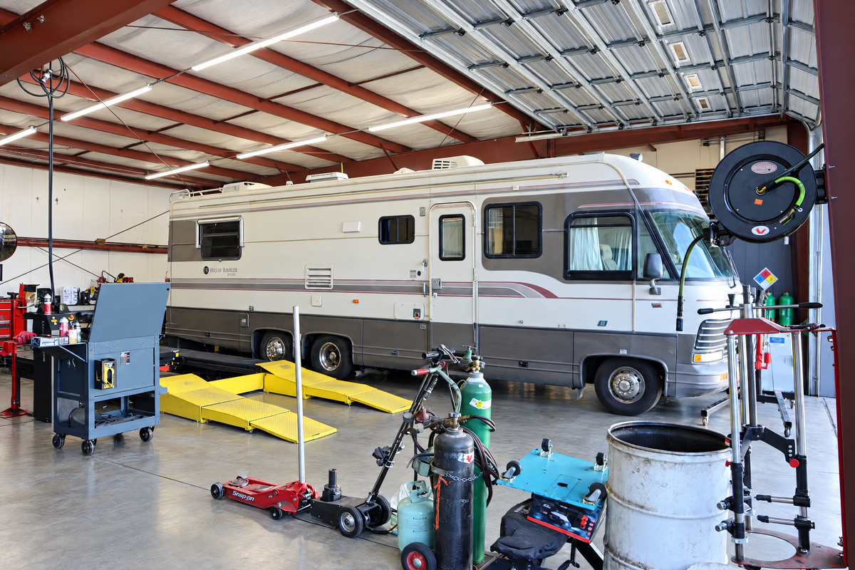 An older Class A motorhome parked inside a shop bay, with welding tanks, a floor jack and drive-on ramps in front of it